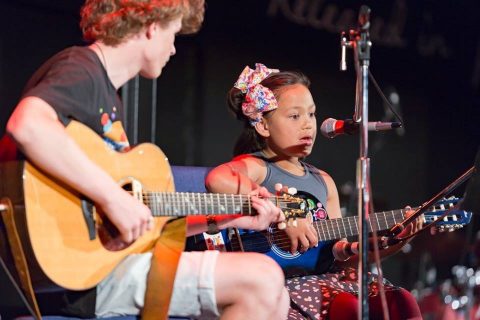 young girl singing and playing guitar with instructor on stage